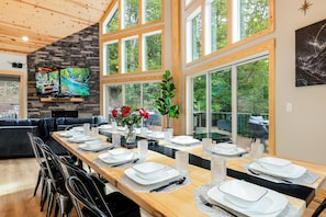 Un comedor con una larga mesa de madera dispuesta para una comida, una chimenea de piedra y ventanas amplias que ofrecen vistas al exterior.
