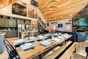 Un comedor espacioso con una larga mesa de madera dispuesta para una comida, una cocina con electrodomésticos modernos y una sala de estar acogedora con chimenea.