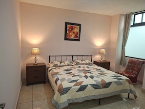 Un dormitorio con una cama, dos mesitas de noche con lámparas, una silla, una ventana con cortinas y un cuadro en la pared.