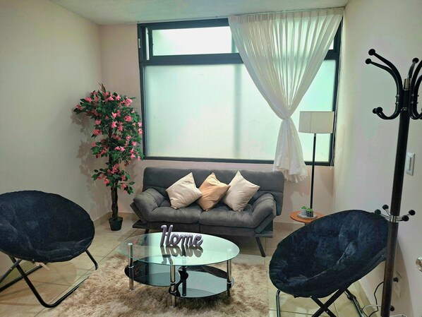 Una sala de estar con un sofá gris, dos sillas negras, una mesa de centro de vidrio, una planta en maceta y una ventana con cortinas.