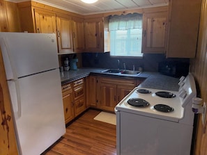 A compact kitchen with wooden cabinets, a white refrigerator, a stove, and a sink. - Grass Lake Hideaway (Gladwin)