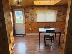 Una habitación con piso de madera, una puerta blanca, una ventana con persianas, una mesa con una silla y un ventilador de techo.