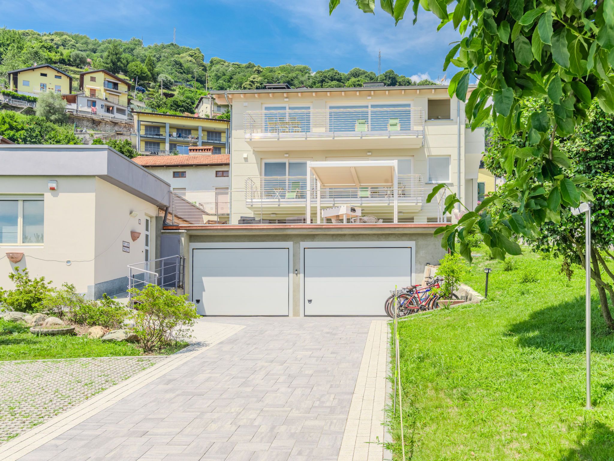 A modern two-story building with a garage, a covered outdoor seating area, and a view of a hillside with other buildings.