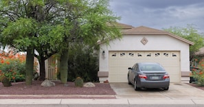 A residential garage with a car parked inside, surrounded by a landscaped garden with cacti and flowering plants. - 3-bedroom house in fantastic East Tucson with cool AC and comfort (Tucson)