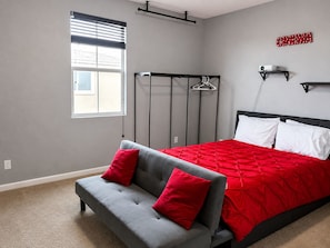 Una camera da letto con un letto, un divano grigio, una coperta a letto rossa e nera, una finestra con persiane e un cartello al muro con la scritta 'CINEMA'.