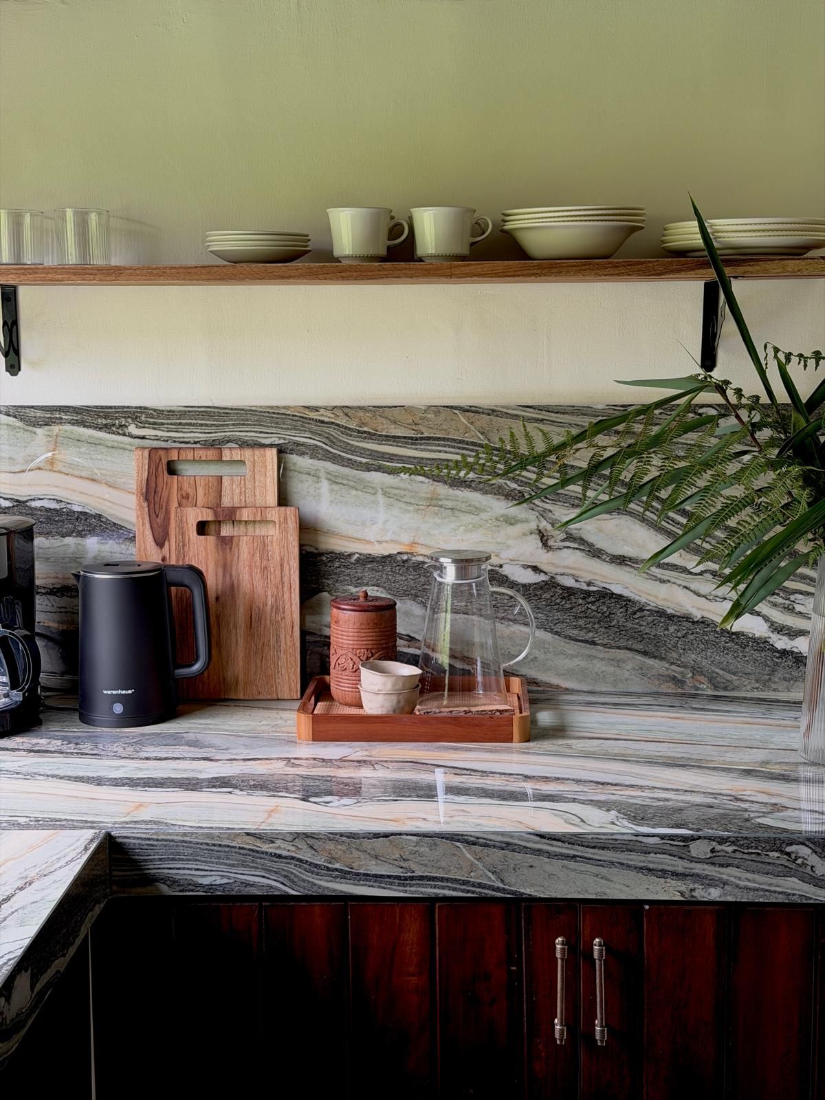 A kitchen with a marble countertop, a coffee maker, a kettle, a wooden cutting board, and a plant.