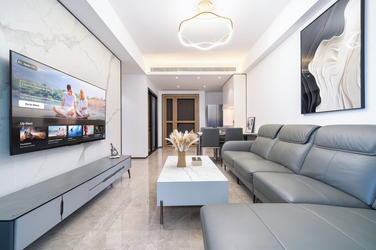 Ein modernes Wohnzimmer mit Flachbildfernseher an der Wand, einem grauen Sofa, einem Couchtisch und einer minimalistischen Einrichtung.