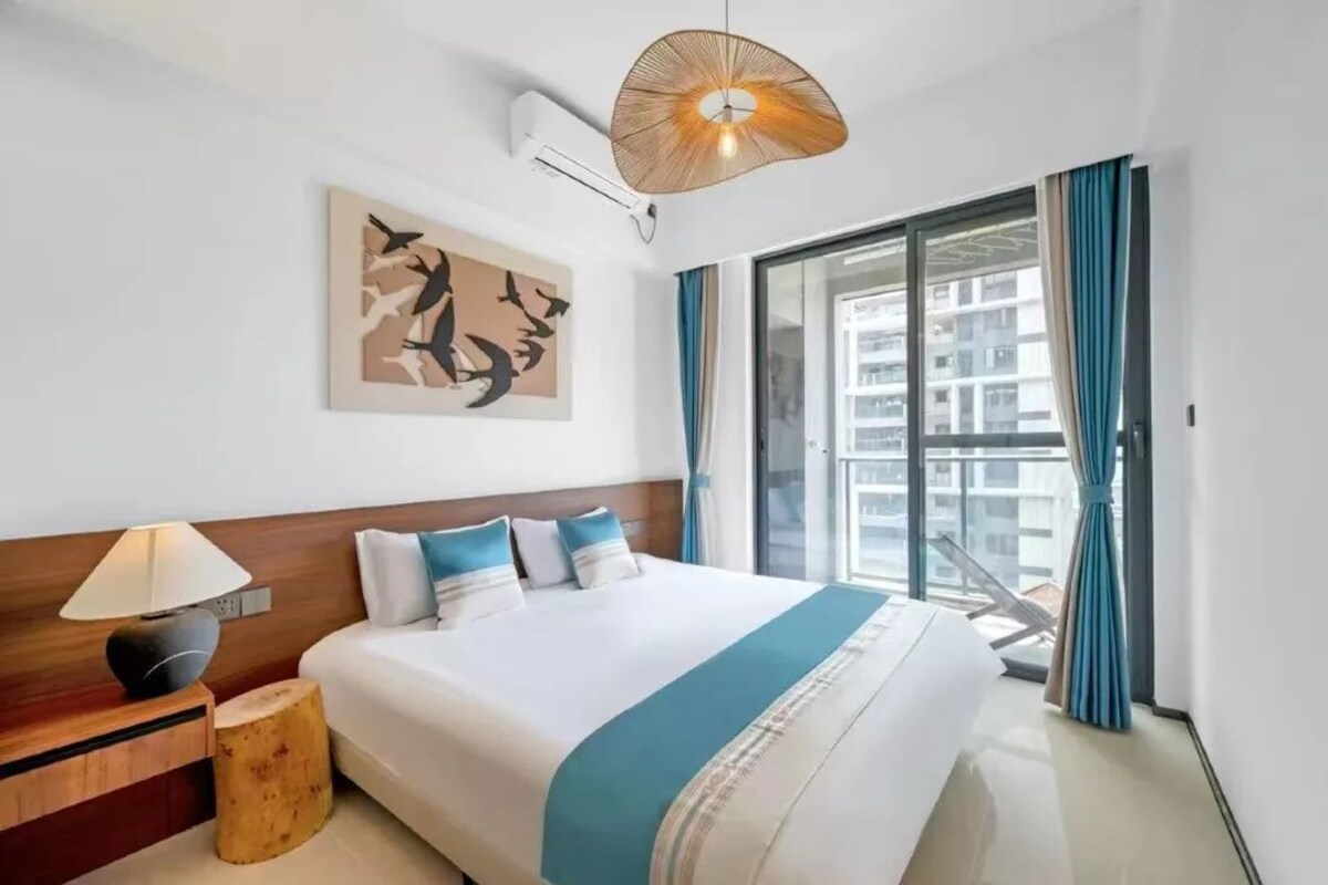 Ein Hotelzimmer mit einem großen Bett, einer Nachttischlampe, einer Wandklimaanlage, einer Deckenleuchte und Blick auf ein Gebäude durch ein Fenster.