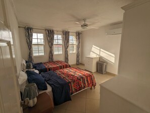 A bedroom with two beds, a ceiling fan, a window with curtains, a small table, and a trash bin. - Kite Surfing, Beach opp Fantastic 1-bedroom apartment with AC . (Oistins)