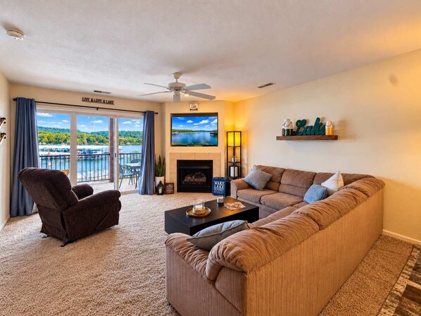 Una sala de estar con sofá, sillón y chimenea. Hay un televisor montado en la pared y un balcón con vistas al lago.
