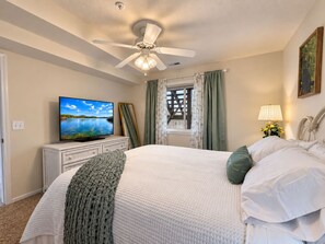 Un dormitorio con cama, ventilador de techo, televisor sobre un aparador y ventana con cortinas.