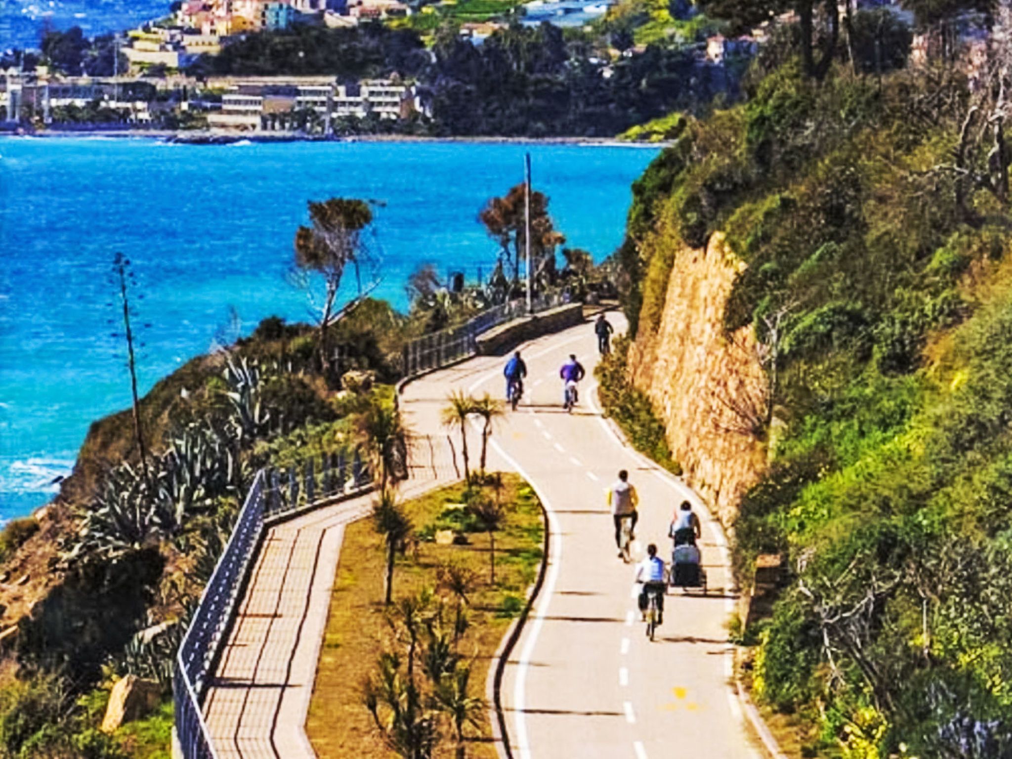A coastal road with cyclists, overlooking a body of water and a hillside with vegetation.