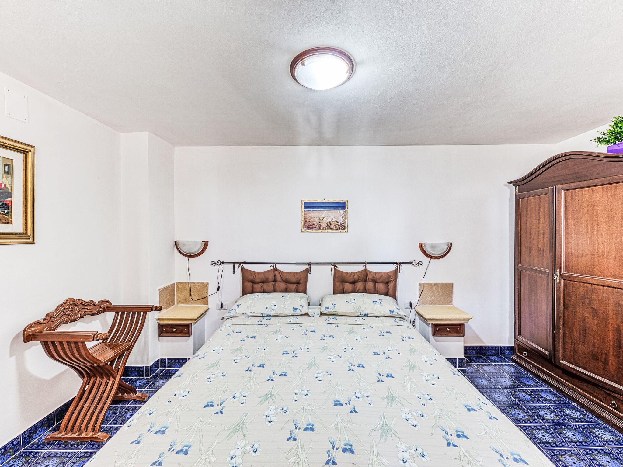 Un dormitorio con cama, mesitas de noche, una silla de madera, un armario y una lámpara de techo.