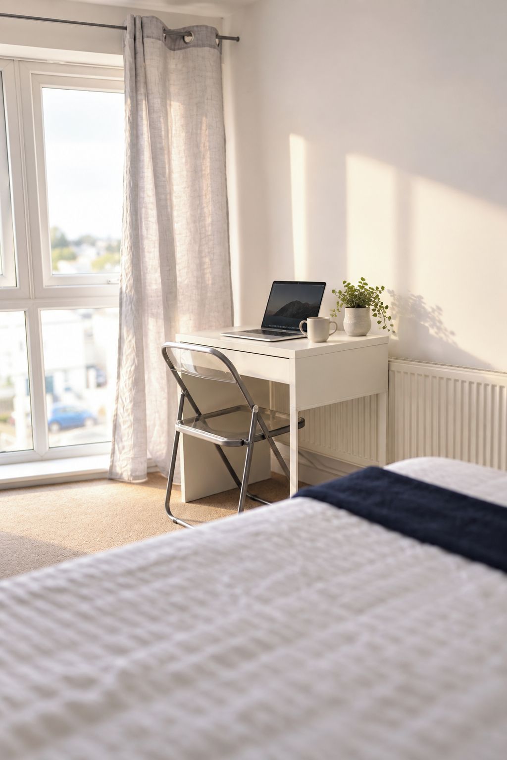A bedroom with a bed, a desk with a laptop, a chair, a window with curtains, and a radiator.