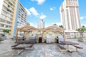 An outdoor patio area with multiple barbecue grills, picnic tables, and a pergola structure. - Stunning 43rd-Fl Studio with Canal Views (Honolulu)