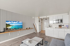 A modern living room with a flat-screen TV, wooden flooring, and a kitchen with white cabinets and stainless steel appliances. - Stunning 43rd-Fl Studio with Canal Views (Honolulu)