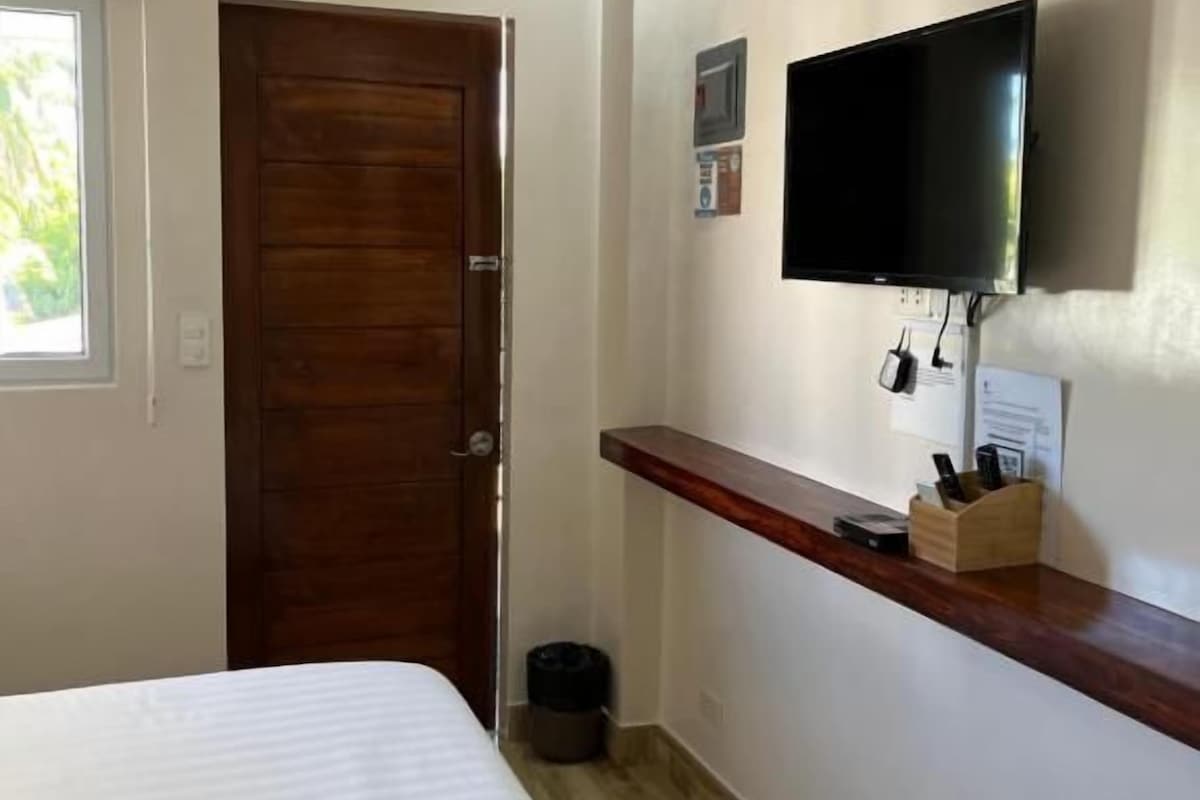 A hotel room with a bed, a wooden door, a television mounted on the wall, and a small shelf with items.