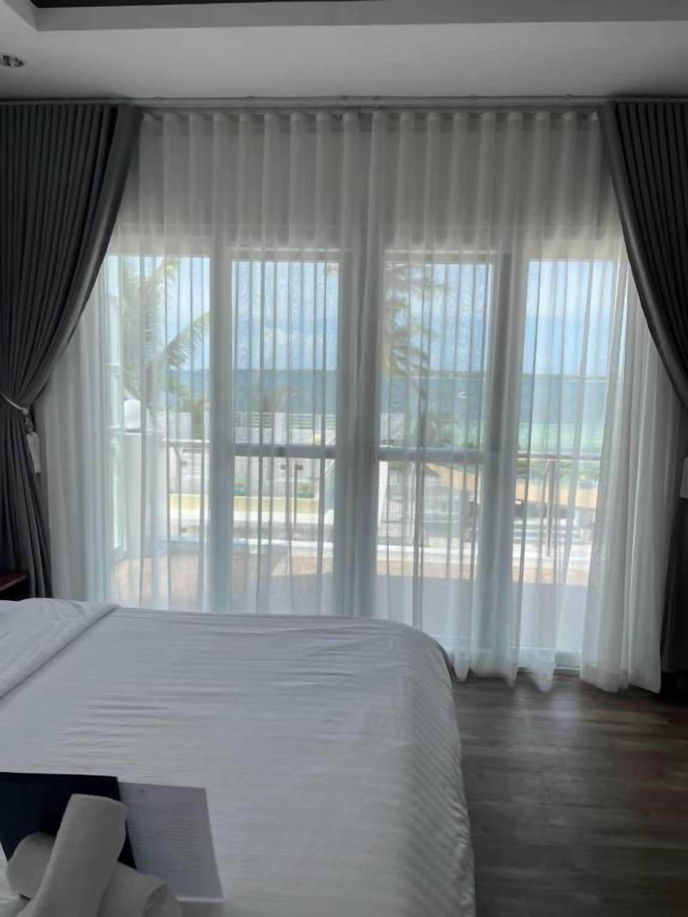 Habitación de hotel con una cama grande, vistas al mar a través de cortinas translúcidas y un balcón con mobiliario exterior.