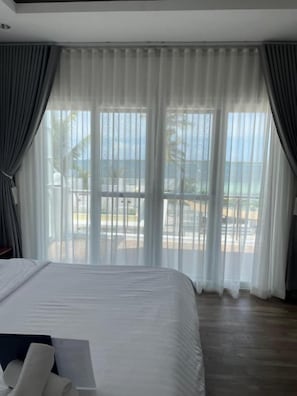 A hotel room with a large bed, a view of the sea through sheer curtains, and a balcony with outdoor furniture.