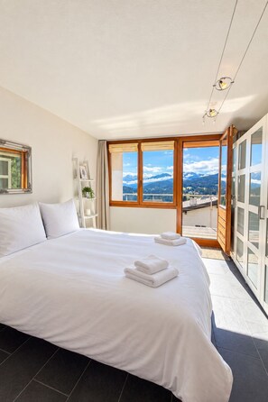 A hotel room with a large bed, a view of mountains, and a balcony with glass doors. - 1511 Foppa 26 by Interhome (Valbella)