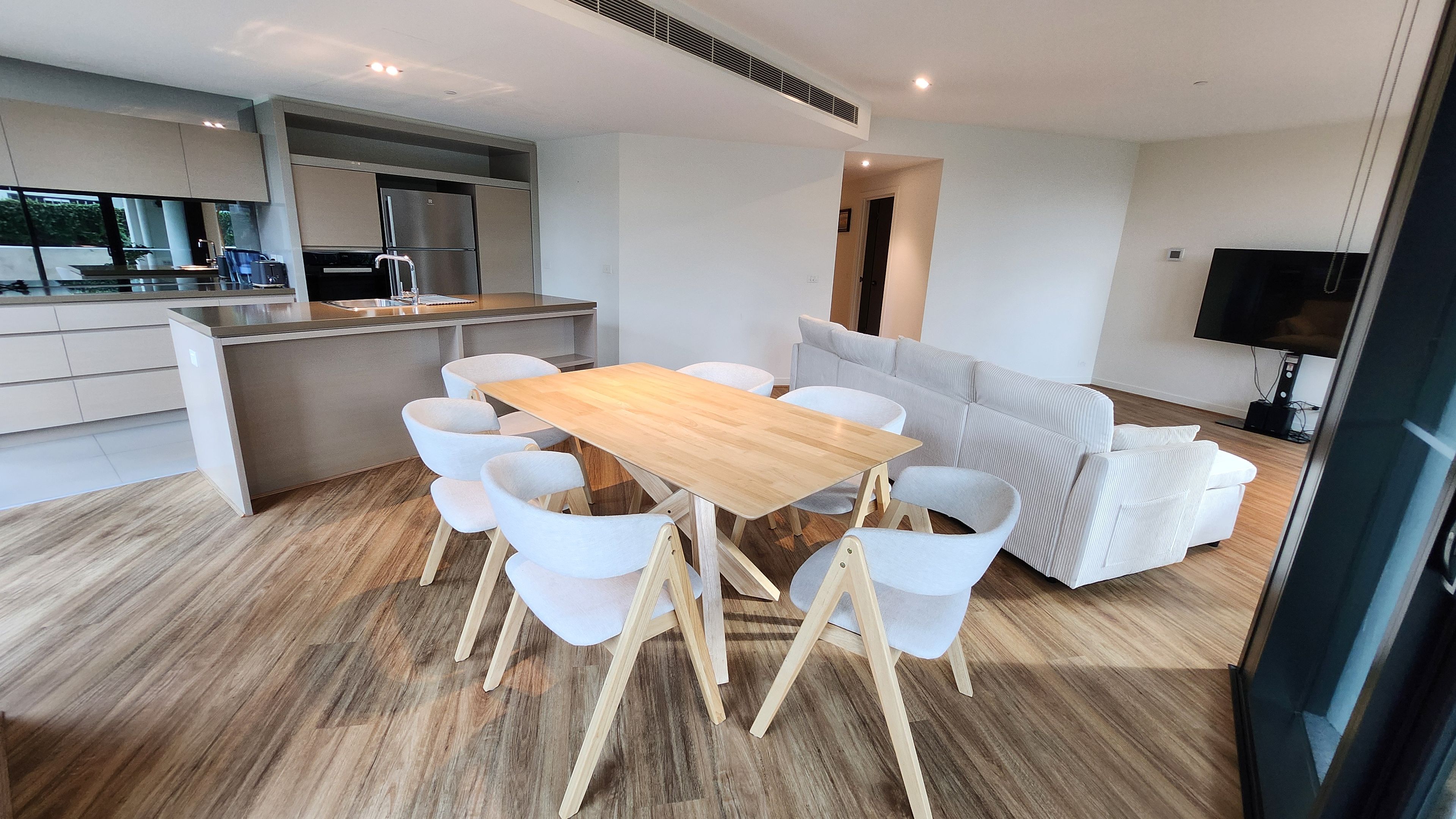 A modern kitchen with a built-in refrigerator and stove, a dining area with a wooden table and white chairs, and a living area with a sofa and television.