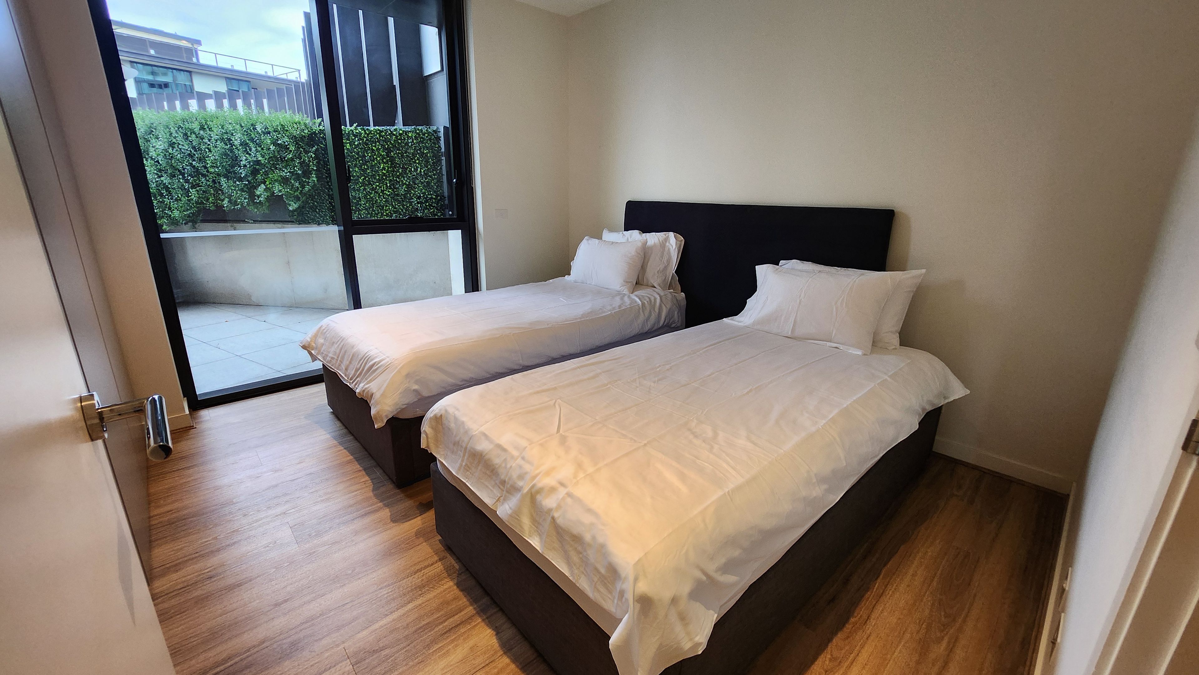 A room with two beds, a sliding glass door leading to a balcony, and a wooden floor.