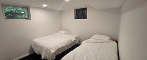 Une petite chambre propre, avec deux lits simples, une fenêtre donnant sur un espace verdoyant et une petite fenêtre avec un rideau.