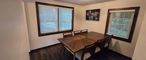 A dining area with a wooden table and chairs, two windows with blinds, and a framed picture on the wall. - Wilder Retreat Lodge near Letchworth - Private Pond, 5BR, Sleeps 10 (Warsaw)