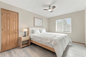 A bedroom with a wooden door, a bedside table with a lamp, a bed with white bedding, a ceiling fan, a framed artwork on the wall, and a window with blinds. - Prime Bellevue Home / Mins to Offutt AFB & Zoo (Bellevue)