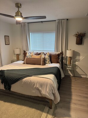 A bedroom with a bed, a ceiling fan, a window with curtains, a bedside table with a lamp, and a wall-mounted plant. - Chic 2-Bedroom Home with Heated Pool + Bar close to AMI/IMG (Bradenton)