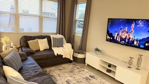 A modern living room with a dark brown leather sofa, a white console table, and a flat-screen TV displaying a castle scene. - The Magic House, Near Disney, Game Room, 10 Guests (Kissimmee)