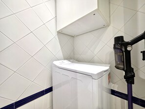 A white washing machine in a corner with a blue stripe on the floor, a black vacuum cleaner on the right, and white tiled walls. - SR Romana Playa 811, directly on the beach (Marbella)
