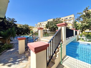Área da piscina com escadaria que leva a um edifício, cercada por vegetação e um céu azul límpido.