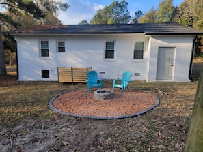 A white building with a brick exterior, a front door, and a window. There is a circular fire pit area with two chairs and a bench. - Charming 3-bedroom house in Southern Pines (Southern Pines)