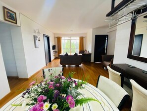 A modern living room with a sofa, a dining table with a floral centerpiece, and a large window with curtains.