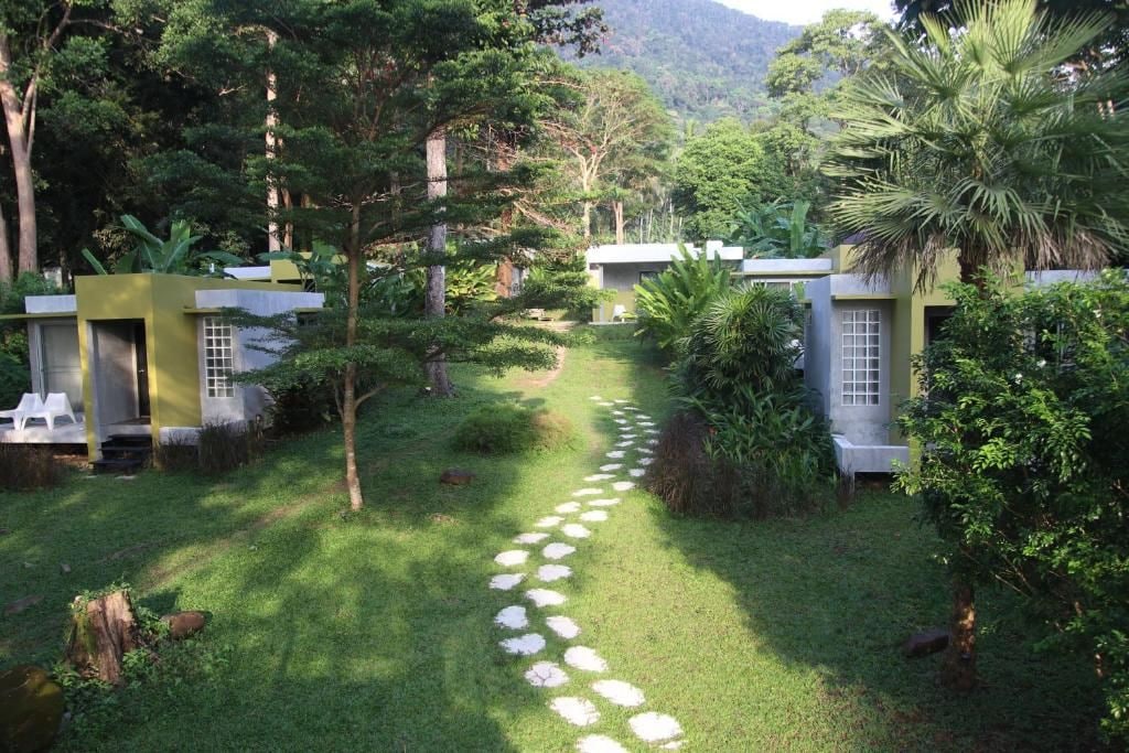 Un sendero atraviesa un jardín exuberante con construcciones pequeñas, rodeado de abundante follaje y árboles.