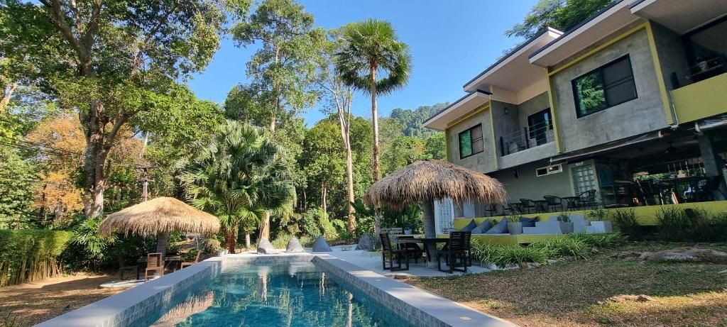 A swimming pool with a thatched roof structure, surrounded by lush greenery and a building with outdoor seating.