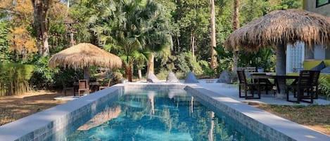 A swimming pool with a thatched roof structure, surrounded by greenery and a dining area with chairs.