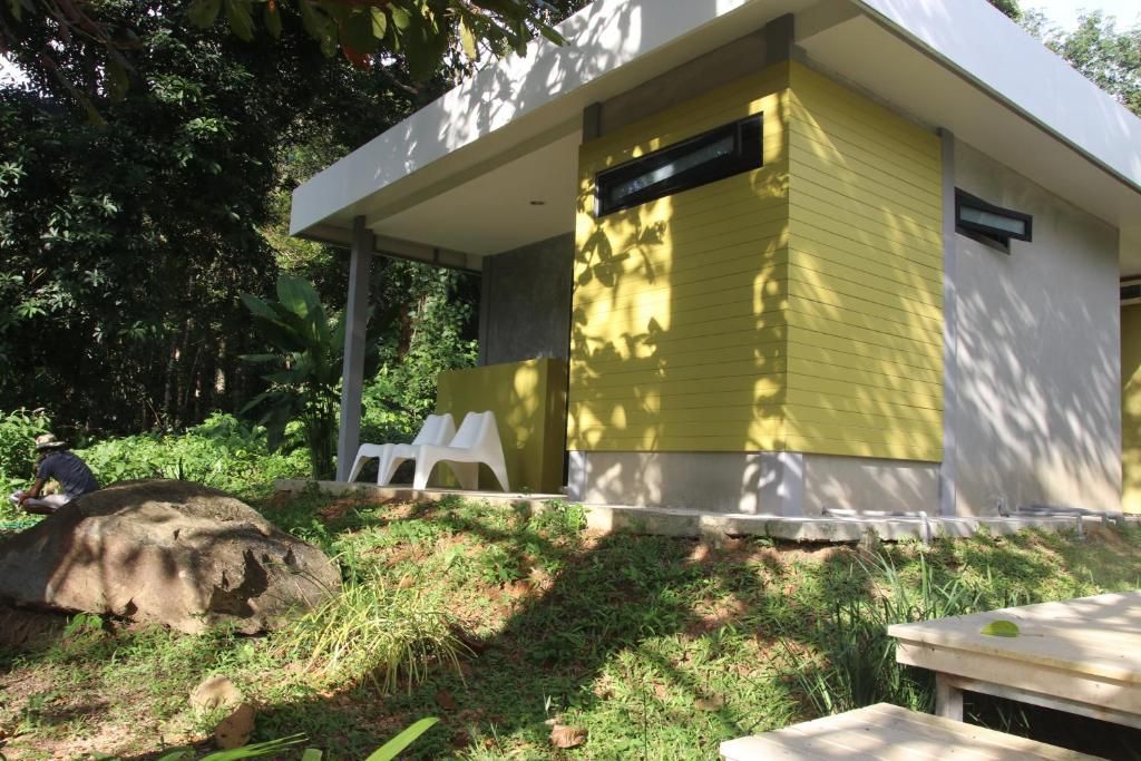 A modern building with a yellow exterior, a covered outdoor seating area with white plastic chairs, and a natural setting with trees and vegetation.