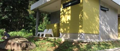 A modern building with a yellow exterior, a covered outdoor seating area with white plastic chairs, and a natural setting with trees and vegetation.