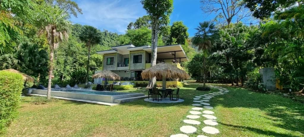 Uma casa de dois andares com piscina, cercada por vegetação exuberante e um gazebo com telhado de palha.