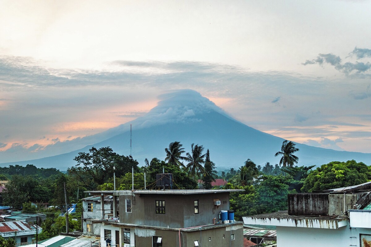 A view of a volcano with a clear sky, surrounded by residential buildings and palm trees.