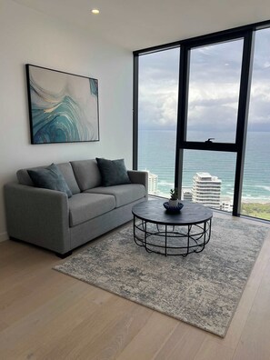 A modern living room with a gray sofa, a round coffee table, and a large window overlooking the ocean. - Panoramic Ocean Luxe Lv54  2Bed 2Bath Parking Inc (Broadbeach)