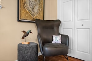Un sillón de cuero negro con un cojín, un taburete de madera con una escultura de pájaro y una obra de arte enmarcada en la pared.