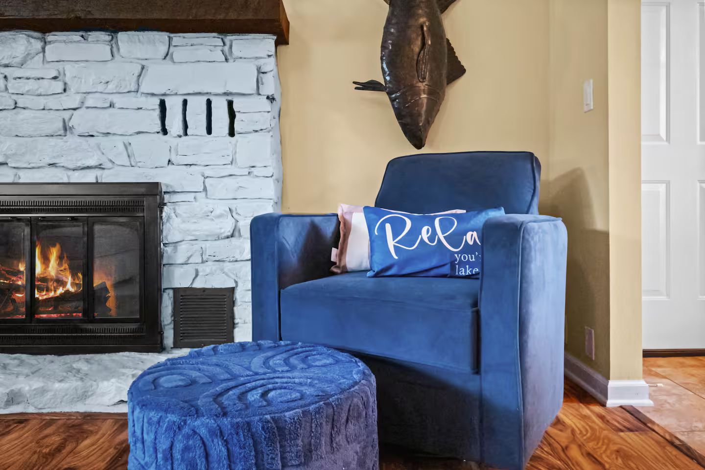 Una habitación con chimenea de piedra, un sillón azul con un cojín y un otoman redondo.