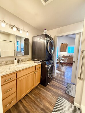 A modern kitchen with a built-in washer and dryer, wooden cabinets, a granite countertop, and a view into the dining area. - The Poulsbo Cottage - Walk to Historic Downtown (Poulsbo)