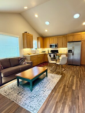 A modern kitchen with wooden cabinets, a stainless steel refrigerator, and a dining area with a table and chairs. - The Poulsbo Cottage - Walk to Historic Downtown (Poulsbo)