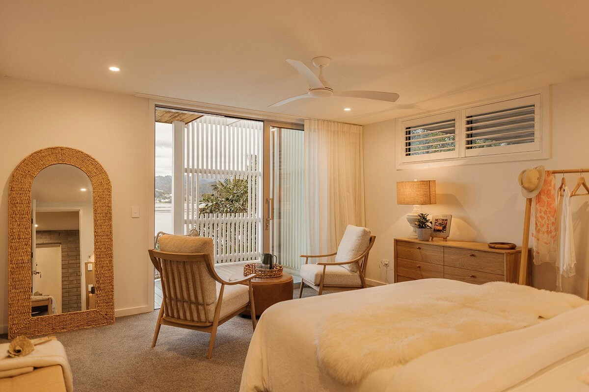 A hotel room with a large bed, two chairs, a small table, a mirror, a ceiling fan, and a view of the outdoors through a sliding door.