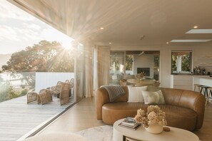 A modern living room with a leather sofa, a round coffee table, and wicker chairs. There is a glass door leading to an outdoor deck. - Twin Tides (Coromandel)