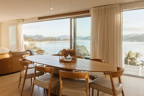 A dining area with a wooden table and chairs, a view of a body of water, and a balcony with a view of the landscape. - Twin Tides (Coromandel)